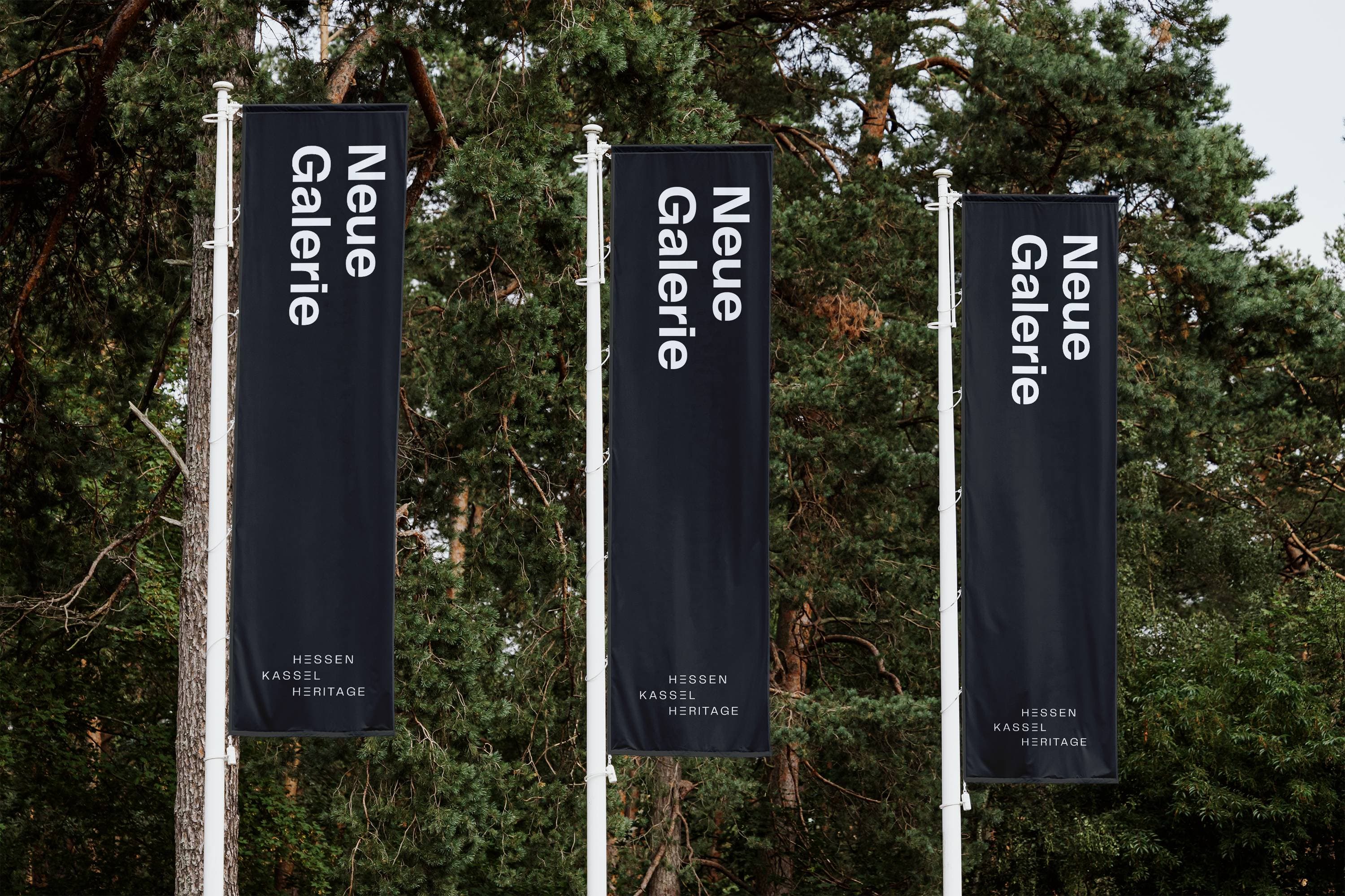 Vor dem Hintergrund hoher grüner Bäume stehen drei vertikale Banner mit der Aufschrift „Neue Galerie“.