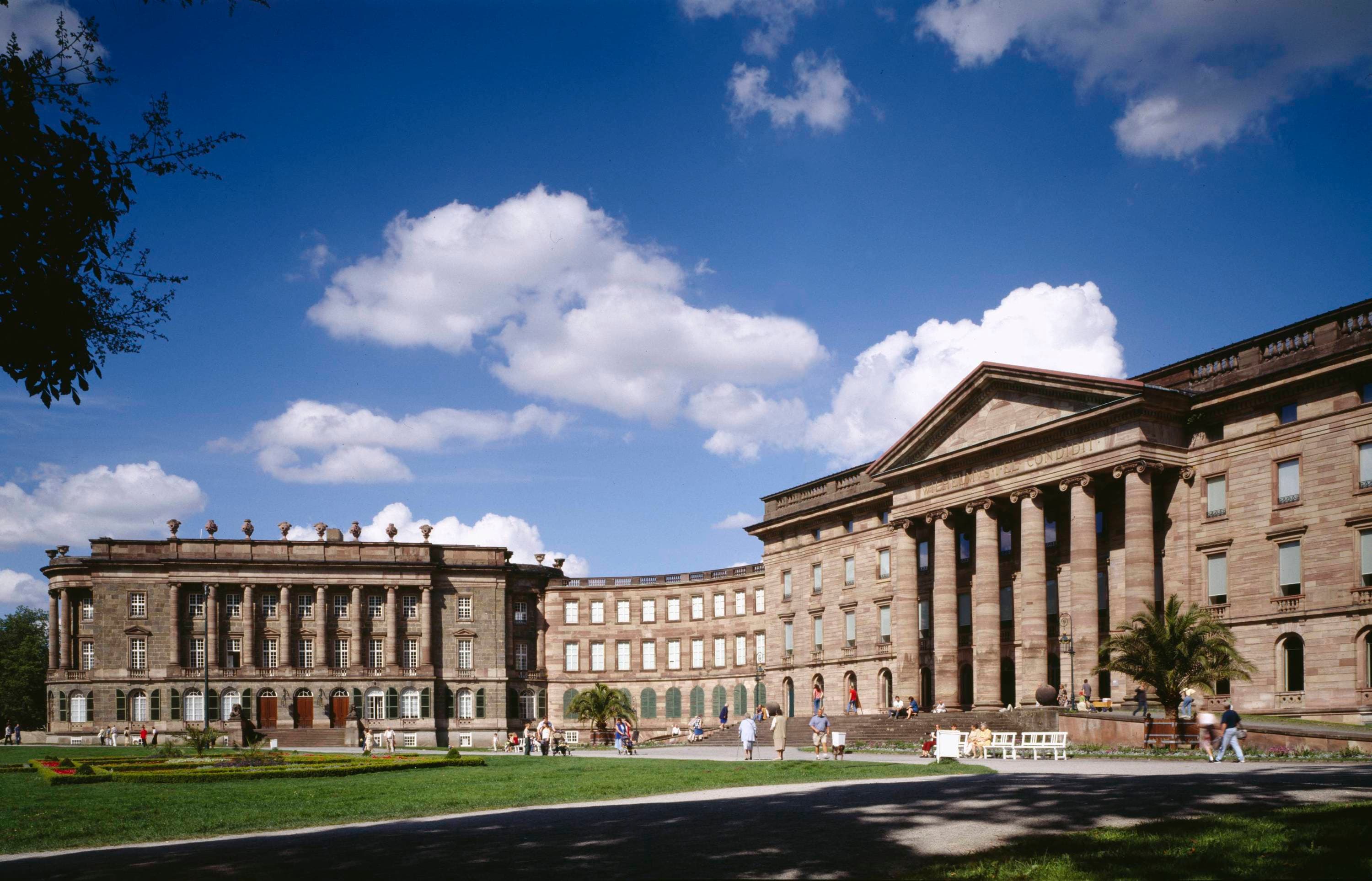 Ein großes neoklassizistisches Gebäude mit Säulen und geschwungener Fassade unter einem blauen Himmel mit vereinzelten Wolken. Menschen gehen auf dem Rasen spazieren.