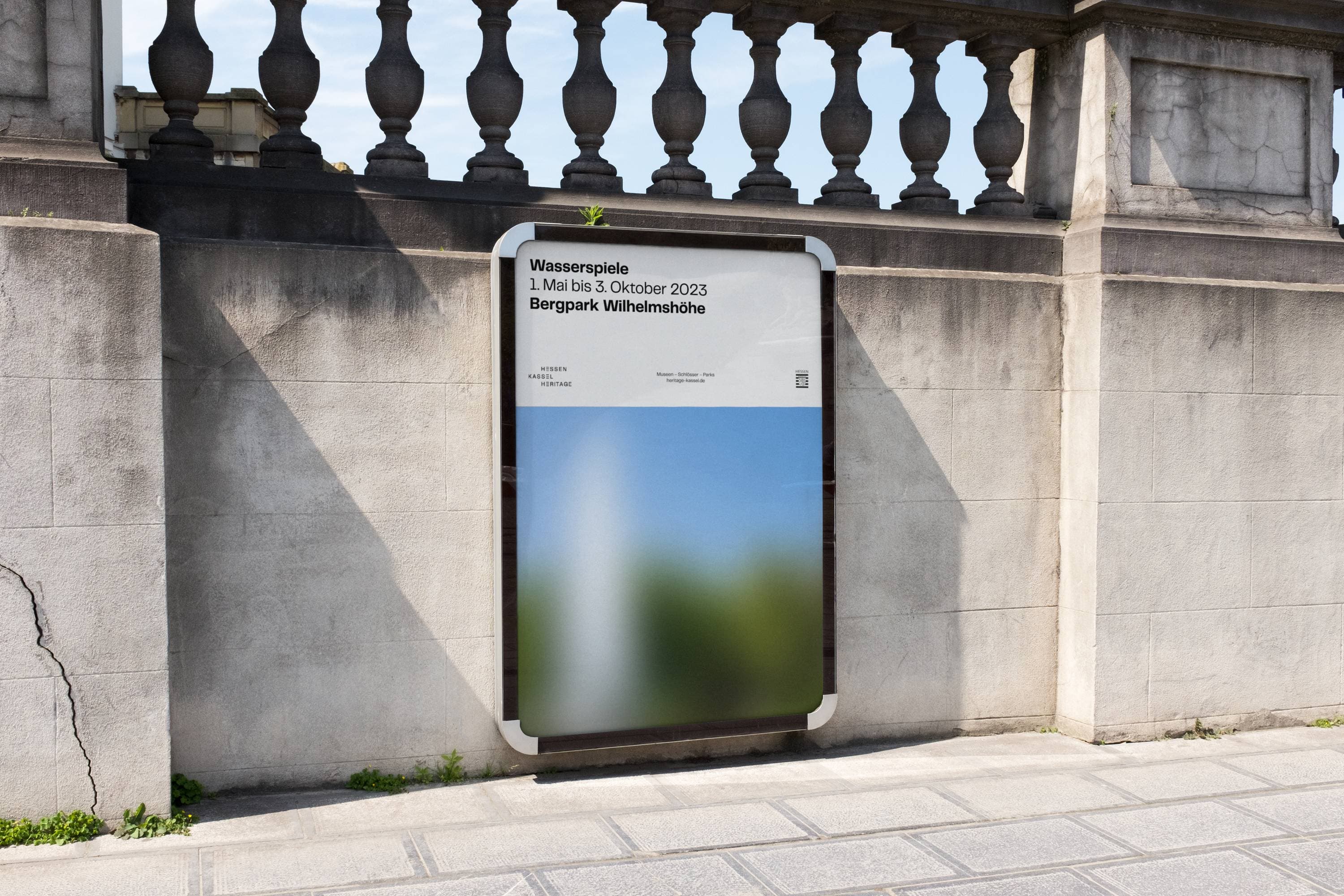 Plakat an einer Steinmauer mit unscharfem Bild, das für die Veranstaltung „Wasserspiele“ vom 1. Mai bis 3. Oktober 2023 im Bergpark Wilhelmshöhe wirbt.